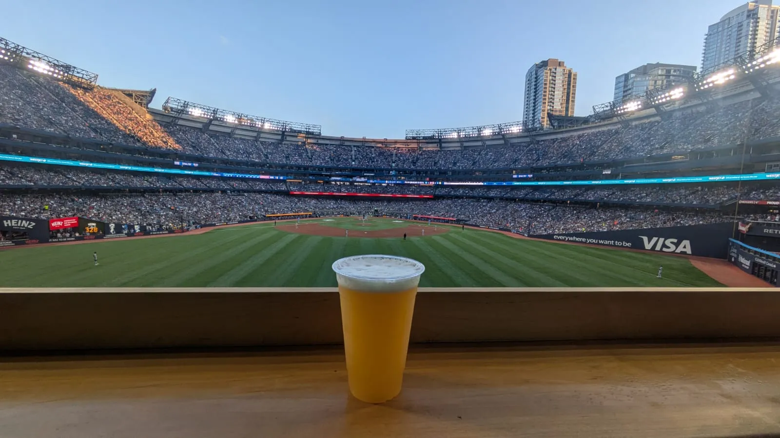 A plastic cup of beer resting on a ledge above the outfield of a packed baseball stadium at dusk.