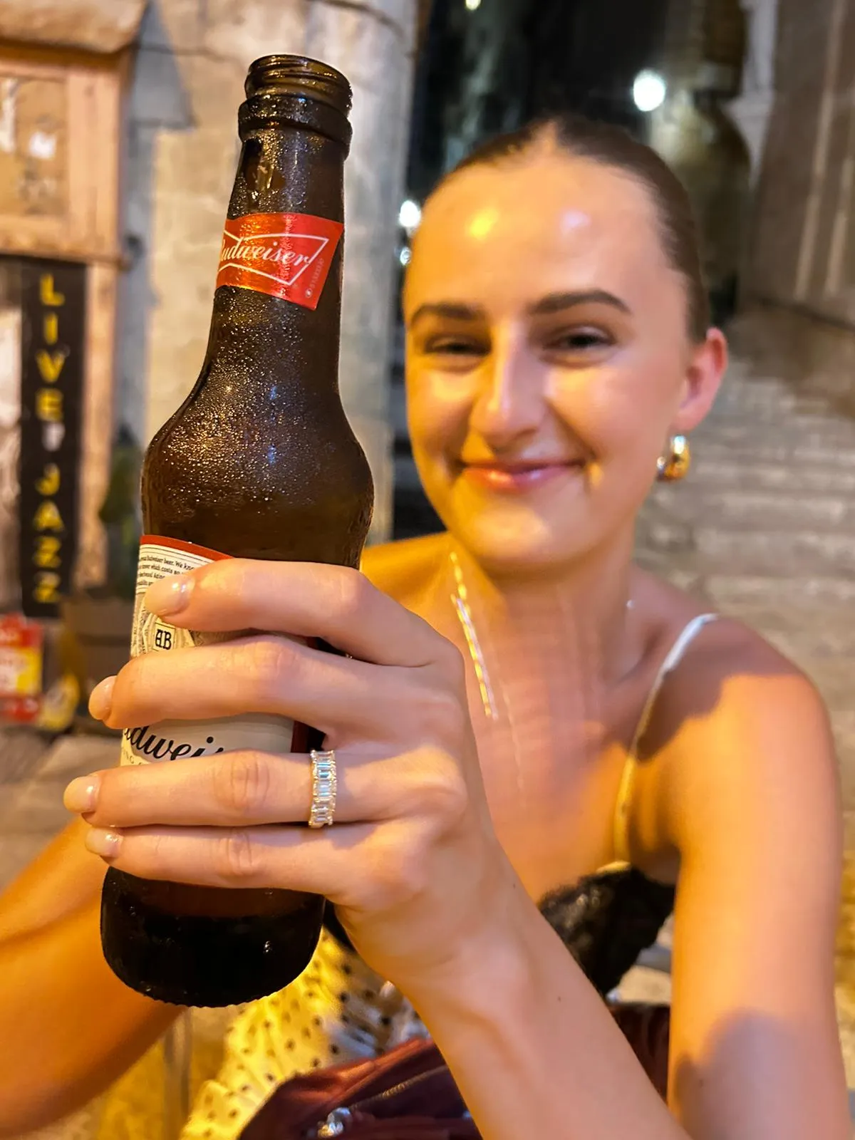 A frosted Budweiser bottle held up to the camera outside a bar, a neon LIVE JAZZ sign glowing in the background.