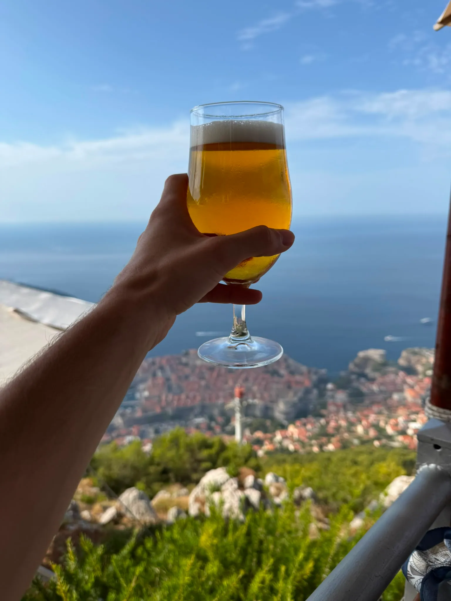 A tulip glass of pale lager held up against a clear sky, high above the red-tiled rooftops of a walled coastal old town with the sea beyond.